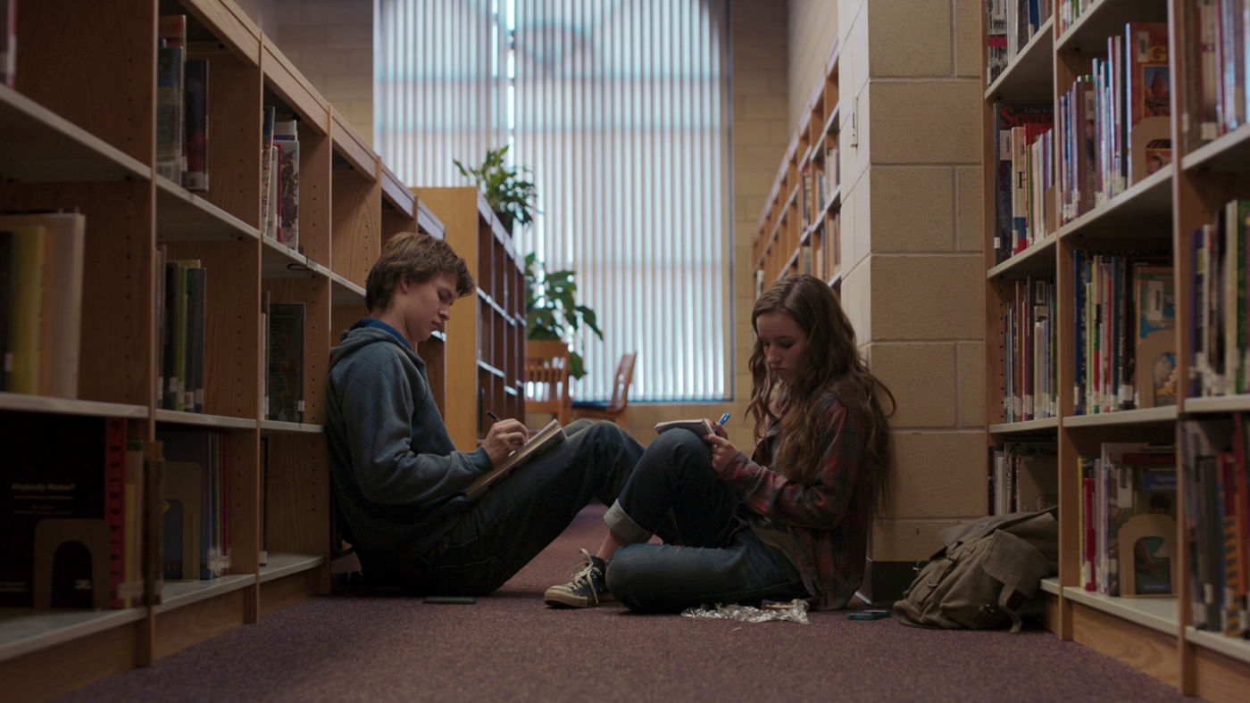 Les deux personnages principaux, assis au sol d’une bibliothèque, entre les étagères de livres, partagent un moment de concentration tandis qu’ils écrivent chacun de leur côté.