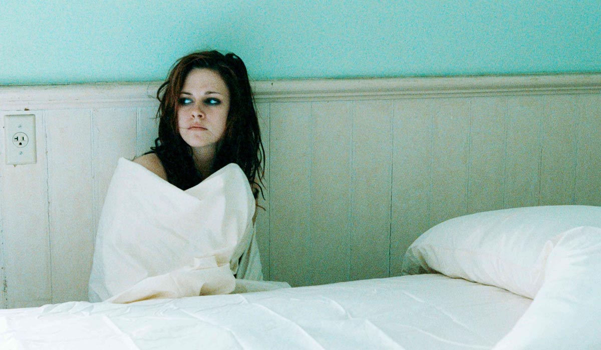 Le personnage de jeune femme incarné par Kristen Stewart, assis derrière un lit, se couvre d’un drap blanc et regarde vers l’extérieur. L’image est claire, ses couleurs pastel. Le regard et les cheveux sombres de la jeune femme contrastent.